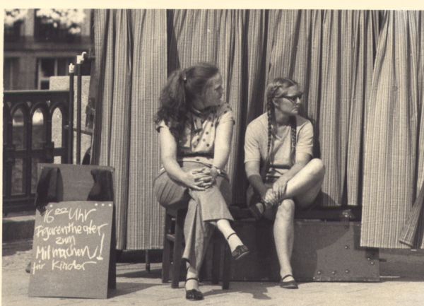 Schwarzweiß Foto zeigt zwei Frauen die vor einem Vorhang auf einem Stuhl und einer Kiste sitzen. Links daneben steht eine Tafel, auf der mit Kreise geschrieben steht „16 Uhr Figurentheater zum Mitmachen für Kinder“