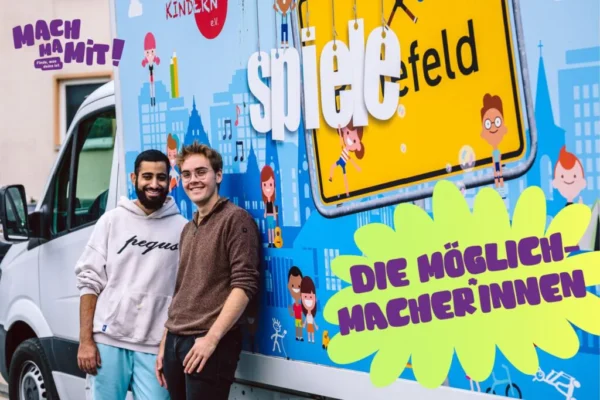 Es sind zwei männlich gelesene Personen zu sehen, die vor einem LKM stehen. Auf dem LKW ist ein Schriftzug Spielefeld zu sehen. Ein Projekt der Kampagne Machmamit. Das Bild ist mit einem Schriftzug die Möglichmacher:innen versehen.