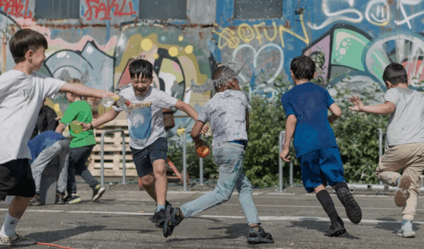 Fünf glückliche Kinder im Sommer rennen auf einem geteerten Platz und machen sich nass mit Wasser.  Im Hintergrund ist eine mit Graffiti besprayte Wand.