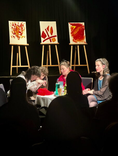 Eine Theaterszene bei der vier Personen an einem niedrigen Tisch vor drei mit Bildern versehenen Staffeleien sitzen. Im Vordergrund sieht man die schwarzen Umrisse dreier Besucher*innen von hinten