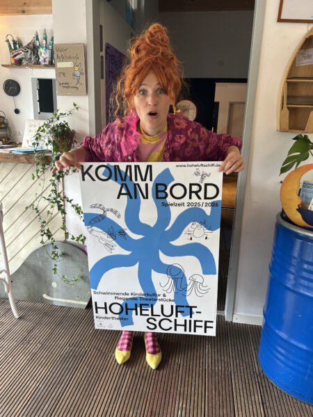 Eine Frau mit rothaariger Turmfrisur steht im Inneren eines Hausbootes mit einem Plakat in der Hand. Auf dem Plakat steht Komm an Bord Hoheluftschiff mit einem hellblauen Fleck, der wie eine Krake aussieht