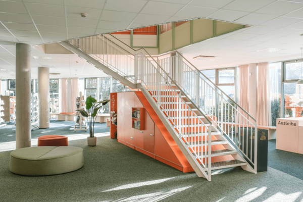 Lichtdurchflutete Innenansicht der Bücherhalle Rahlstedt mit weißem Treppenaufgang über orangefarbenen Schränken, Grünpflanze und Sitzpontons