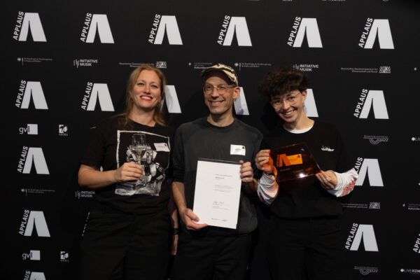 Drei Personen stehen mit Urkunde, Preis und einem Glas Wein vor der Werbewand des APPLAUS Awards