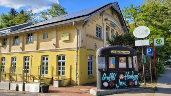 Ein gelbes Guthaus-ähnliches Gebäude im Grünen mit einem Bushalteschild und einem Teil U-Bahn-Waggon als Kunstwerk und Anschlagbrett auf dem Graffiti Hamburg Jam und U5 Kulturinsel steht