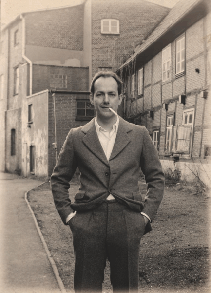 Schwarz-weiß Foto eines Mannes vor einer schiefen Fachwerkhaus-Fassade. Der Mann hat eine Zigarette im Mundwinkel und die Hände in den Hosentaschen seines Anzugs.