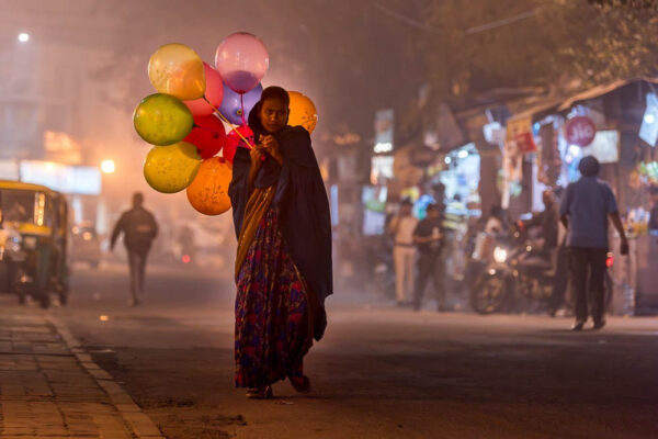 Eine Frau im Sari mit einer bunten Ballontraube geht durch eine nächtlich erleuchtete Straße, auf der verschwommen andere Menschen, Fahrzeuge und Geschäftsfassaden zu sehen sind
