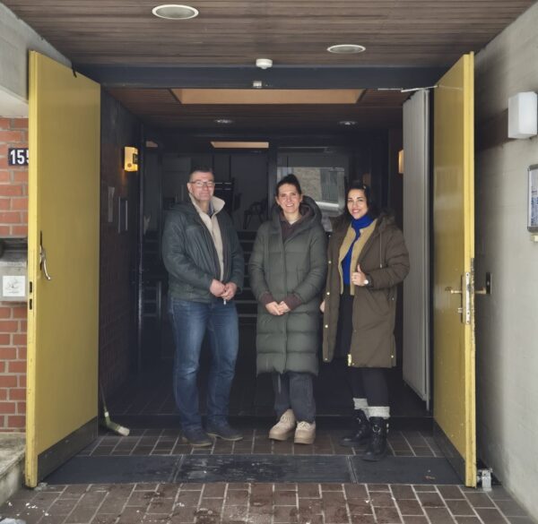 Drei Personen stehen mit Winterjacken in einem Hauseingang, der von gelben Türen flankiert ist.