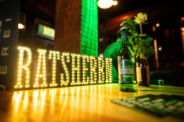 Ein Bierflasche steht zusammen mit einer gelben Rose auf einem Tresen. Im Hintergrund sind ein Kühlschrank, ein menschlicher Rücken und der Leuchtschriftzug Ratsherrn zu sehen
