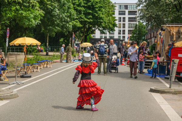 Ein sommerliche Straße mit Passant*innen, Ständen, Bäume und Sitzgelegenheiten. Mitten drin: Ein von hinten fotografiertes Kind im roten Flamencokleid mit silbernem Zylinder