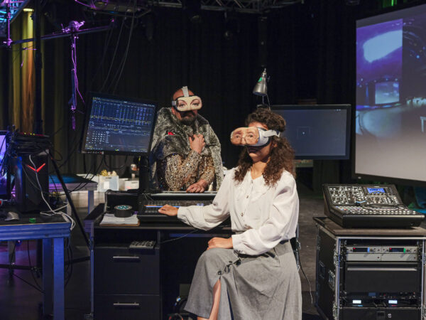 Zwei Personen mit VR-Brillen sitzen vor technischen Geräten in einer Bühnensitzuation