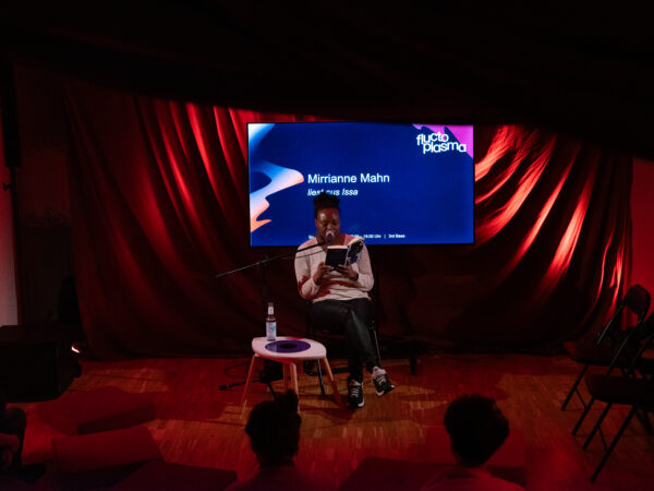 Eine Person sitzt ebenerdig auf einem Stuhl mit einem Buch in der Hand, aus dem sie vorliest. Im Hintergrund zeigt ein Bildschirm das Logo des Fluctoplasma Festivals