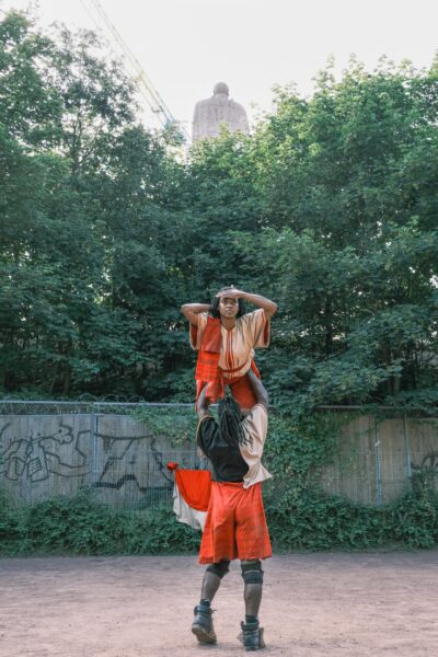 Es sind zwei BIPoC-Personen zu sehen, die eine Hebefigur bilden. Die untere Person steht mit dem Rücken zum Betrachter. Die gehobene Person hält sich die rechte Hand vor die Stirn, die linke Hand hinter den Kopf. Die Personen tragen rot, gold, schwarze Gewänder. Die Personen sind vor dem Bismarck-Denkmal und grünen Bäumen positioniert.