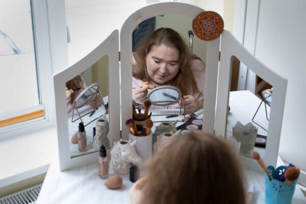 Eine Person mit langen Haaren schminkt sich vor einem weißen Schminktisch. Ihr Gesicht ist durch die Spiegelung fotografiert.