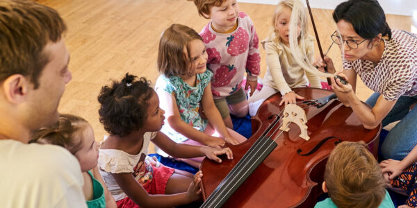 Kinder, die um einen liegenden Kontrabass stehen und knien, dessen Funktion eine Person mit Brille erklärt