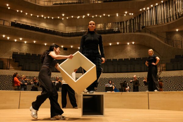 Foto von drei Personen auf der Bühne in der Elbphilharmonie Hamburg, die eine Choreographie mit Kuben aufführen. Im Hintergrund spielt ein mehrköpfiges Streicherensemble