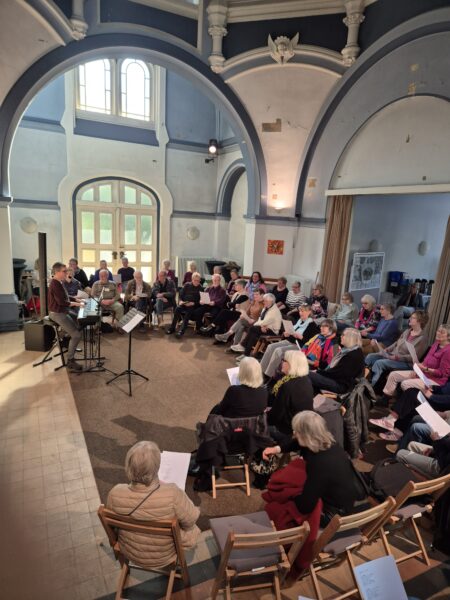Menschen, die im Halbkreis auf Stühlen unter der blau-weißen Kuppel einer Kapelle sitzen und singen bzw. der Musik einer Person an einem Keyboard lauschen.