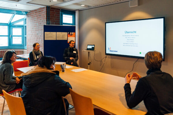Menschen sitzen in einem Raum um einen rechteckigen Tisch und vor einem Smartboard auf dem eine Übersicht angezeigt wird