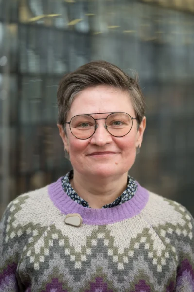 Portrait einer weißen Person mit kurzen Haaren, Brille und einem farbigen Wollpullover, die offen in die Kamera guckt
