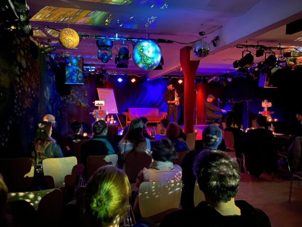 Foto des Saals der Motte mit Bühnenbeleuchtung und Menschen im Publikum, einer Person, die Saxofon spielend auf der Bühne steht. In der Mitte des Raums hängt eine als Welt stilisierte Diskokugel