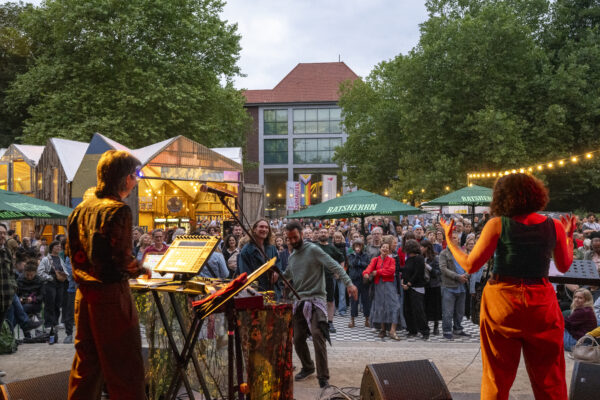 Foto während eines altonale-Konzerts von der Bühne aus mit Blick auf die Rücken der Künstler*innen ins teils tanzende Publikum. Im Hintergrund das Altonaer Theater , Bäume und Buden