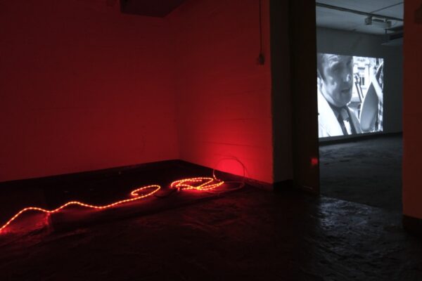 Ein rot gestrichener Raum mit einer rötlich leuchtenden Licherkette am Boden und einem Durchbruch zu einem anderen Raum, in dem auf einer wandhohen Leinwand ein Schwarzweiß-Film gezeigt wird