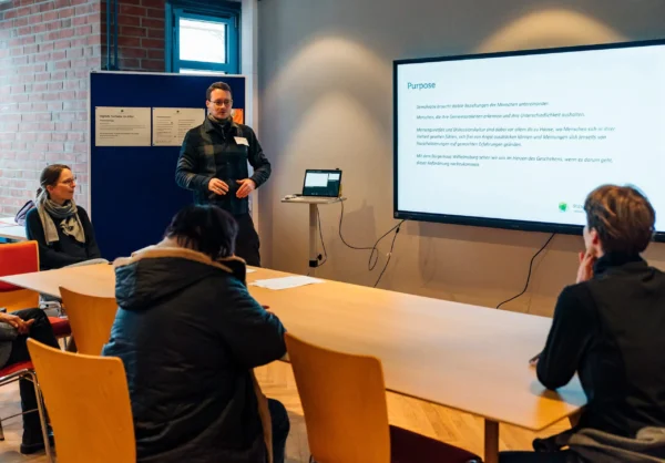Menschen sitzen in einem Raum um einen Tisch mit Blick auf ein Smart Board, vor dem ein Mensch Inhalte des Infotextes, der auf dem Board zu sehen ist, erklärt