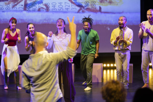 Menschen in einer Bühnensituation, bei einer Probe, von denen eine Person im Vordergrund steht und singt, während jemand sie mit erhobener Hand mit Victory-Zeichen animiert