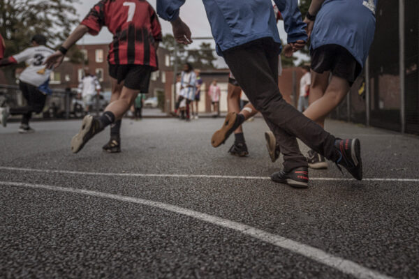 fünf Personen von hinten auf Asphalt fotografiert, die jeweils ein Bein hinter das andere gekreuzt haben, wie im schwungvollen Tanz. Drei Personen tragen ein Fußball-Trikot und Shorts