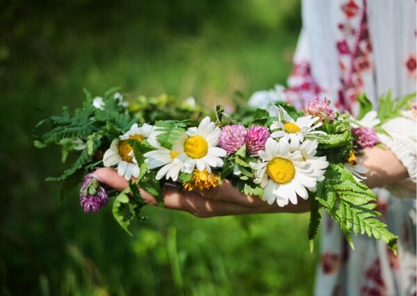 Ein Blütenkranz mit rosa und weiß-gelben Blumen und grünem Farn, auf den Händen einer Person, die verschwommen am Bildrand auf einer grünen Wiese zu erahnen ist